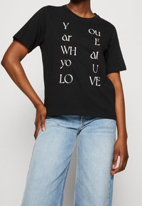 Camiseta de algodón negra con texto vertical blanco: "Eres quien amas." Con mangas cortas y un ajuste relajado.