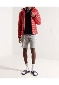 Rote Steppjacke, schwarzes T-Shirt, graue Shorts, weiße Socken und marineblaue Slide-Sandalen mit Logo. Schlichter Hintergrund mit strukturierter Wand.