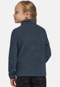 Bambino con capelli intrecciati, che indossa una giacca in pile blu scuro e pantaloni neri, rivolto di lato su uno sfondo neutro.