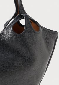 Fekete bőrtáska, texturált felülettel, kivágott fogantyúkkal és kontrasztos barna belső résszel. Görbült forma, minimális részletezéssel.