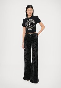 Črna kratka majica z logotipom, kombinirana s črnimi žametnimi hlačami na zvonec z zapletenim damastnim vzorcem in dvema gumboma spredaj.