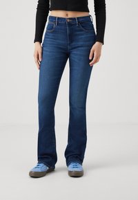 Tummansiniset farkut, joissa on istuva vyötärö, levenevät lahkeet ja kontrastiväriset ompeleet. Yhdistetty vaaleansiinisiin tennareihin, joissa on sileä denim-rakenne.