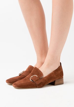 Mocassins en daim marron avec des détails de boucle en or portés pieds nus, présentant un petit talon empilé sur un fond blanc.