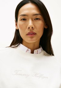 Felpa crema con tessuto testurizzato, logo ricamato "Tommy Hilfiger", indossata sopra una camicia a righe rosa chiaro con colletto.