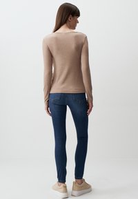 Maglietta beige a maniche lunghe con scollatura a U, abbinata a jeans skinny blu scuro e sneakers beige, vista da dietro. Texture liscia.