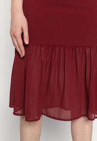 Vestido burgundy com um corpo justo e uma bainha plissada, fluida e transparente. A textura é suave, com um material leve e fluido.