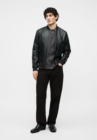 Νέος άντρας που φοράει μαύρο δερμάτινο μπουφάν, πουκάμισο με σχέδια σε σκούρο χρώμα, μαύρο παντελόνι και μαύρα παπούτσια, στέκεται με το ένα χέρι στην τσέπη.