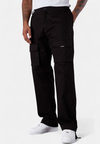 Schwarze Cargo-Hosen aus strapazierfähigem Material, mit mehreren Seitentaschen, einem lässigen Schnitt und einem Reißverschlussdetail in der Tasche. Getragen mit weißen Sneakers.