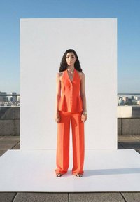 Oranžna brezrokavna peplum majica s sprednjimi gumbi, v kombinaciji z širokimi hlačami. Gladek material, oblečeno z črnimi sandali, postavljeno pred belo ozadje.