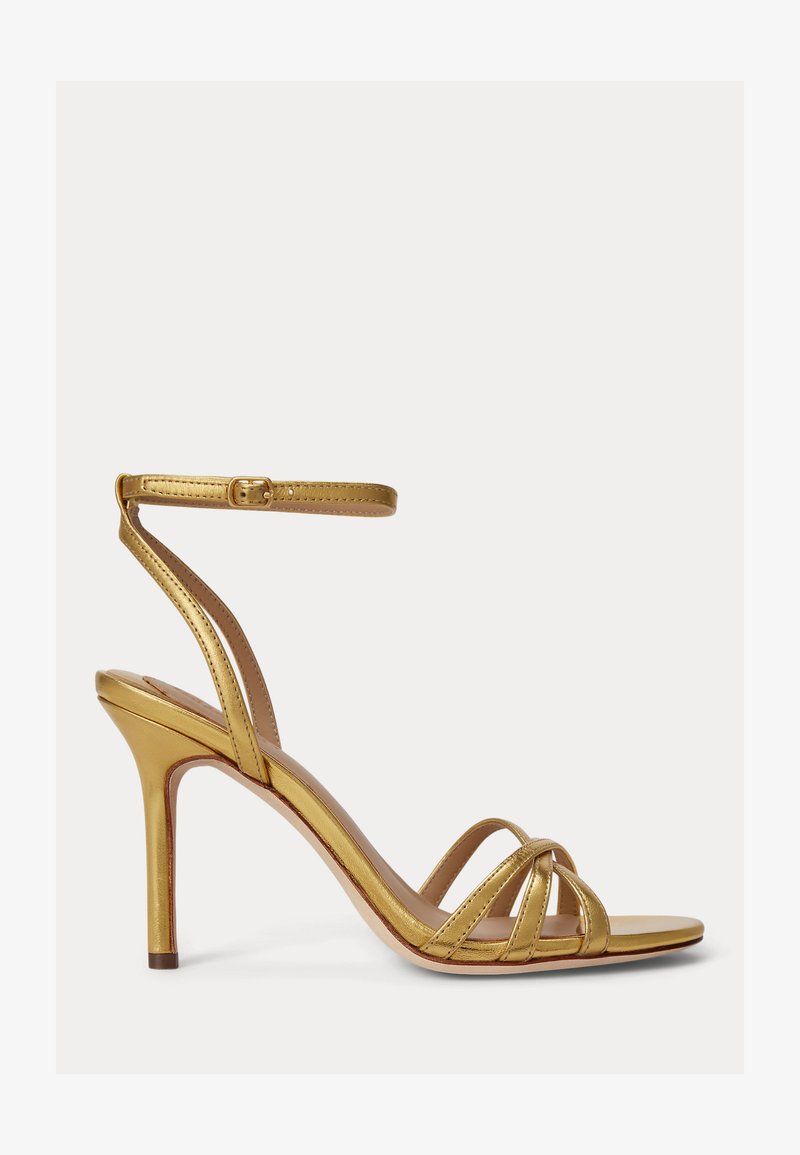 Χρυσό μεταλλικό σανδάλι με ψηλό τακούνι, λεπτές λουρίδες πάνω από τα δάχτυλα και λουράκι αστραγάλου με πόρπη, σε λευκό φόντο.