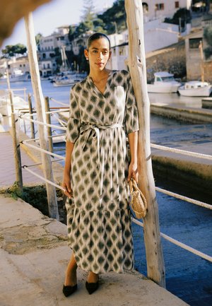 Kobieta w długiej sukience w wzory i czarnych butach, trzymająca plecioną torbę, stoi przy drewnianym słupie blisko portu z łodziami i budynkami w tle.