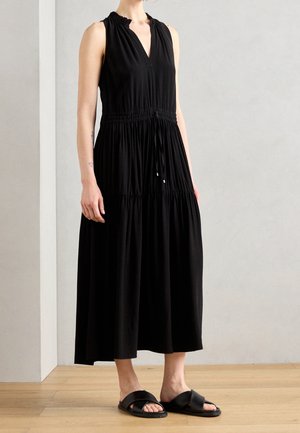 Personne portant une robe midi noire sans manches avec taille froncée et des sandales noires à enfiler, debout sur un sol en bois contre un mur uni.