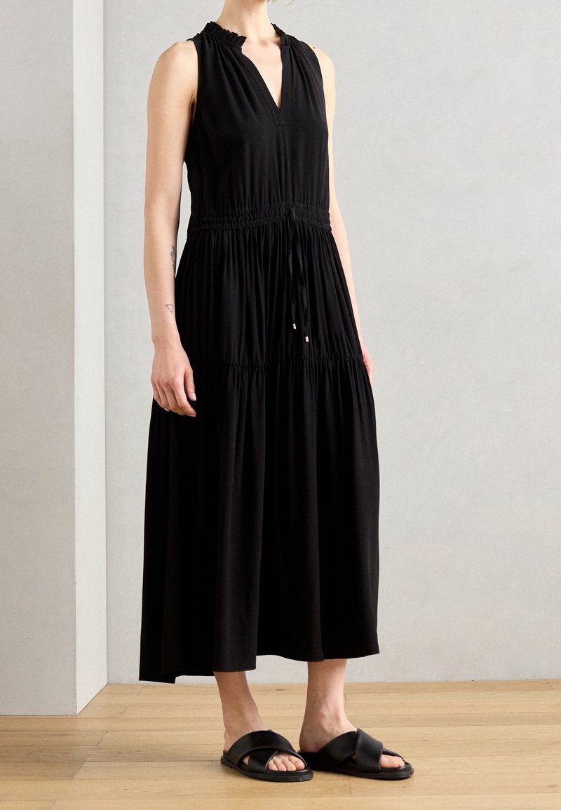 Personne portant une robe midi noire sans manches avec taille froncée et des sandales noires à enfiler, debout sur un sol en bois contre un mur uni.