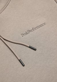 Beige hoodie med dragsnören som har svarta spetsar, broderad "Peak Performance"-logotyp på framsidan, tillverkad av strukturerad tyg.