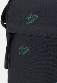 Borsa in tessuto nero con texture, decorata con due loghi di coccodrillo ricamati in verde e una tracolla nera regolabile con un cursore in plastica.