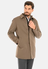 Cappotto marrone impermeabile con collo a risvolto, bottoni frontali e tasche laterali, indossato sopra un maglione grigio e una camicia bianca.