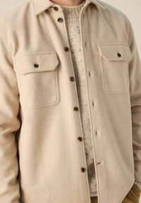 Veste en mélange de laine beige avec deux poches poitrine, boutons marron foncé, et un pull crème texturé en dessous.
