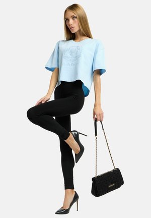 Hellblaues, kurz geschnittenes T-Shirt mit kurzen Ärmeln, schwarze Leggings, schwarze High Heels und eine strukturierte schwarze Handtasche mit goldener Kettenriemen.