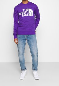 Fialová mikina s bílým logem, kulatým výstřihem a žebrovanými manžetami, doplněná světle modrými džínami a bílými teniskami.