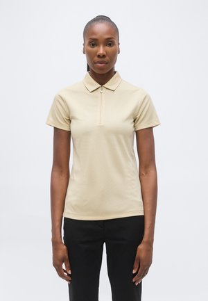 Tummempi-ihoinen nainen, jonka tukka on sidottu taakse, pukeutuneena beigeen lyhythihaiseen vetoketjulliseen poolopaitaan ja mustiin housuihin vaaleaa yksiväristä taustaa vasten.