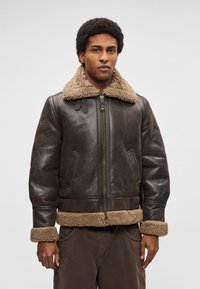Barna bőr bomber dzseki, világosbarna gyapjú gallérral és mandzsettával, cipzáros záródással és oldalzsebekkel. Texturált és tartós kialakítás.