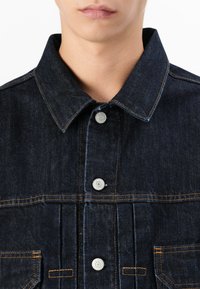 Donkerkleurige spijkerjas met een puntkraag, zilveren knoopsluitingen, oranje stiksels en voorzakken met stikselaccenten.