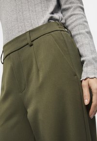 Olivově zelené kalhoty volného střihu, s bočními kapsami a stahovacími šňůrkami v pase, spárované s světle šedým pleteným topem s texturovaným vzorem.