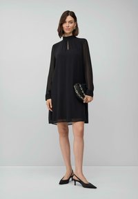 Vrouw in een zwarte lange jurk tot op de knie, met een kleine zwarte handtas met een gouden ketting en zwarte slingback hakken.