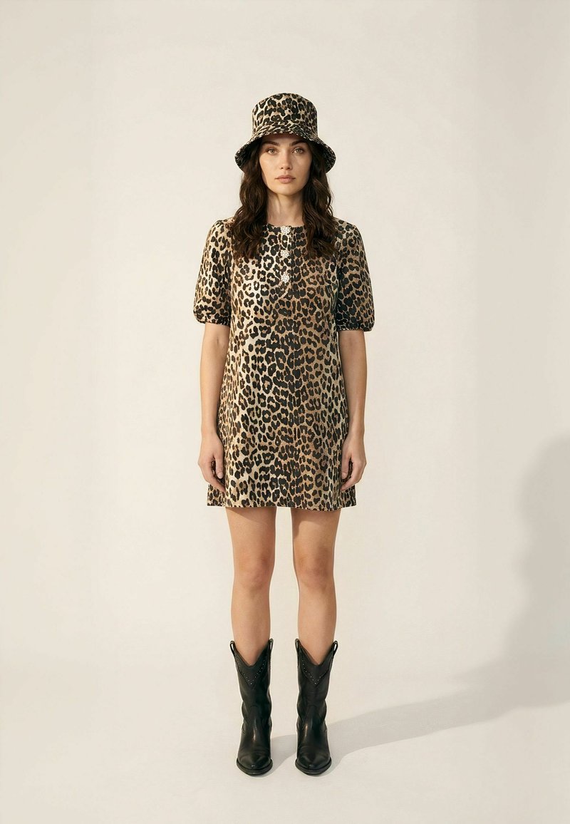 Femme portant une robe à imprimé léopard et un chapeau bob assorti, associée à des bottes de cowboy noires mi-mollet, debout devant un fond uni.