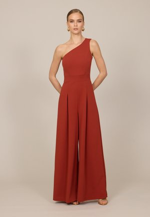 Femme debout portant une combinaison sans manches, une épaule, couleur rouille avec jambes larges, boucles d'oreilles créoles dorées et sandales.