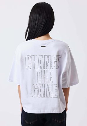 Persona con capelli lunghi neri indossa una maglietta bianca larga con la scritta traforata "CHANGE THE GAME" sul retro.