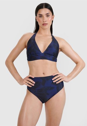 Kobieta o długich ciemnych włosach, ubrana w granatowy kwiecisty top bikini na szyję i majtki z wysokim stanem, z rękami na biodrach na jednolitym tle.