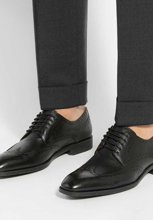 Chaussures habillées en cuir noir de style brogue portées avec un pantalon gris foncé retroussé, laissant les chevilles visibles sans chaussettes.