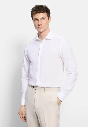 Uomo che indossa una camicia bianca aderente a maniche lunghe e pantaloni beige chiaro, in piedi con una mano in tasca.