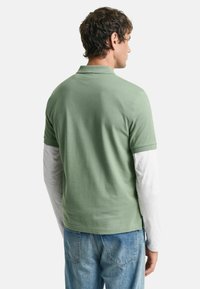 Polo verde de manga corta hecho de algodón, combinado con una capa blanca de manga larga debajo. Ajuste casual con vista desde la parte posterior.