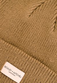 Beżowa, dziana czapka z prążkowanym wzorem i wywiniętym daszkiem, z białą metką z napisem "BICKLEY + MITCHELL" i "AMSTERDAM".