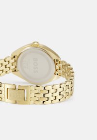 Montre-bracelet dorée avec un bracelet métallique à maillons, un boîtier rectangulaire et une finition polie. Présente un fond solide avec le logo "BOSS".