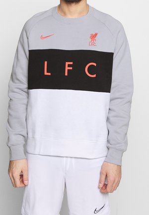 Person, die einen grauen, schwarzen und weißen Sweatshirt mit rotem "LFC"-Text und dem Liverpool FC-Logo trägt, kombiniert mit weißen Nike-Shorts.