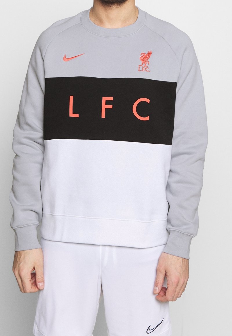 Personne portant un sweat-shirt gris, noir et blanc avec un texte rouge "LFC" et le logo du Liverpool FC, assorti à un short Nike blanc.
