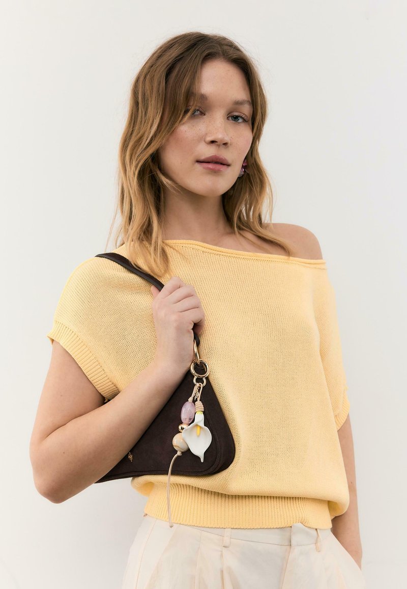 Ung kvinde iført en gul skulderfri strikbluse og cremefarvede bukser, som holder en mørkebrun trekantet håndtaske med dekorative vedhæng.