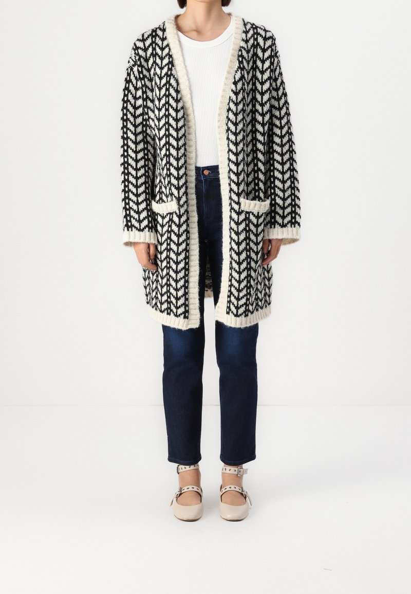 Černobíle vzorovaný kardigan s volným střihem, se vsadkou do V, dvěma předními kapsami a žebrovanými lemy, spárovaný s modrými džíny a světlou obuví.