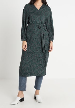Long green dress with a v-neck, floral pattern, belted waist, and long sleeves, paired with light blue jeans and white heeled mules.
