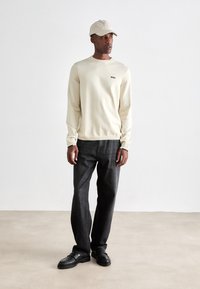Hombre con suéter de cuello redondo beige con el logo "BOSS", gorra beige, jeans gris oscuro y zapatos negros, de pie sobre un suelo claro contra una pared blanca.