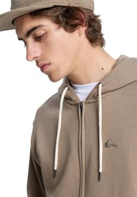Beige zip-up hoodie met een voorlogo, witte trekkoorden en een zachte textuur. Draag het met een crèmekleurig T-shirt eronder.