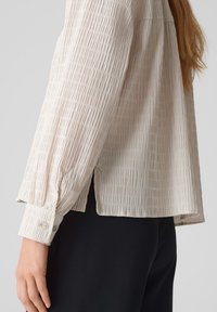 Helle beige, strukturierte Bluse mit vertikalen Falten, langen Ärmeln und geknöpften Manschetten. Über schwarzem Stoff getragen, mit seitlichen Schlitzen.