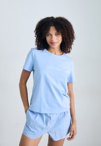 Mladá žena s kudrnatými vlasy se usmívá, má na sobě světle modré tričko Calvin Klein a odpovídající pruhované šortky před jemně šedým pozadím.