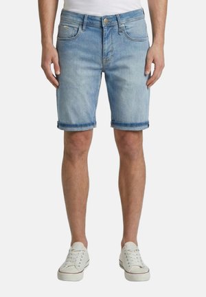 Hombre con pantalones cortos vaqueros azul claro con dobladillo enrollado, zapatillas blancas y una camiseta blanca, de pie frente a un fondo blanco liso.