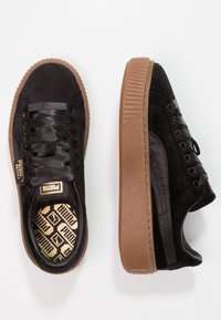 Svarta mockaskor med satinsnören, med en texturerad brun gummisula och guldaccenterad branding inuti skon.