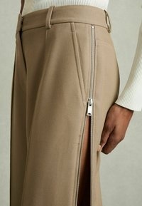 Beige muotit pantssit, joissa on sivuvetoketju ja sivureikä, valmistettu teksturoidusta kankaasta, vyölenkeillä vyötäröllä.