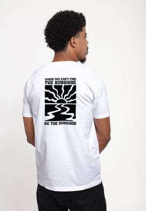 Homme de dos, portant un t-shirt blanc avec un graphique de soleil noir et texte : "When you can't find the sunshine, be the sunshine."